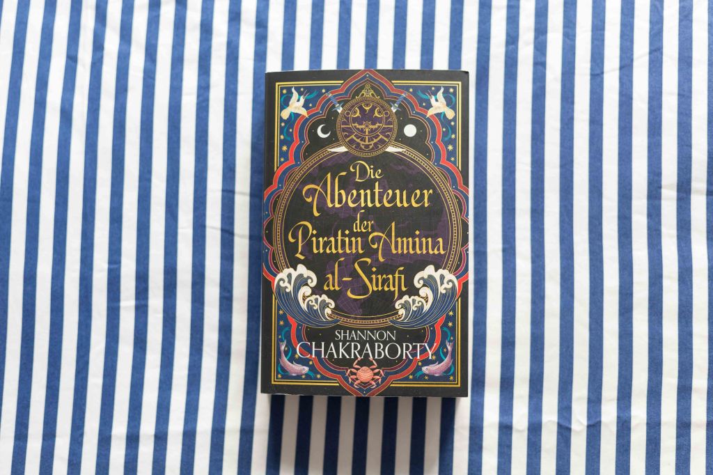 Das Buch liegt auf einem blau-weiß gestreiften Stoff. Das Cover von "Die Abenteuer der Piratin Amina al-Sirafi" zeigt verschiedene Elemente aus Meer und Himmel.