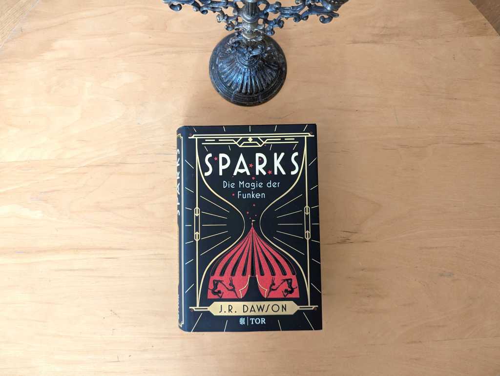 Die gebundene Ausgabe von "Sparks - Die Magie der Funken" liegt auf einem Tisch vor einem Kerzenleuchter. Das Cover zeigt ein rotes Zirkuszelt, in dem die Silhouetten von vier Artistinnen zu sehen sind. Über dem Zelt steigen rote Funken auf.