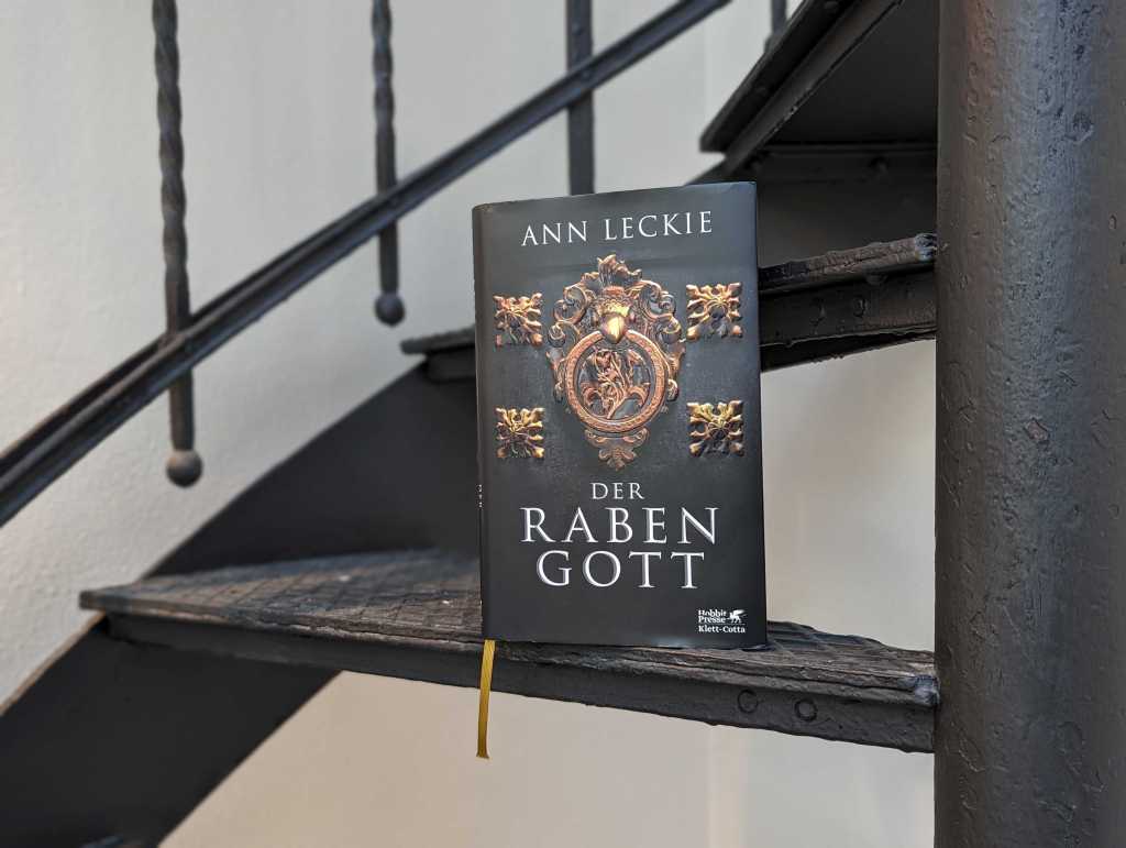 Das Buch "Der Rabengott" von Ann Leckie steht auf einer Treppe. Das Cover zeigt einen mit Ornamenten verzierten goldenen Rabenkopf, der einen Wappenring im Schnabel hält.